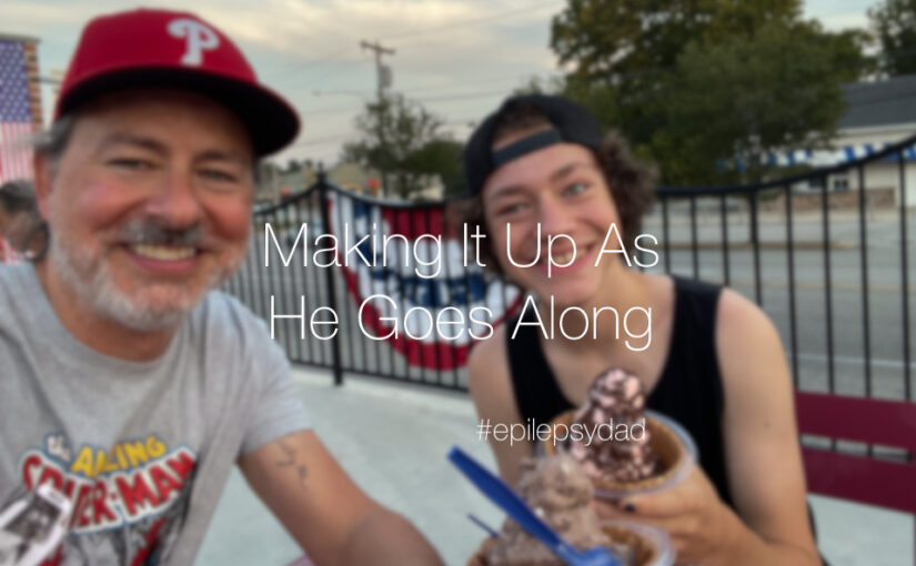 a man in a phillies hat sitting at a table outside with a teenage boy eating ice cream